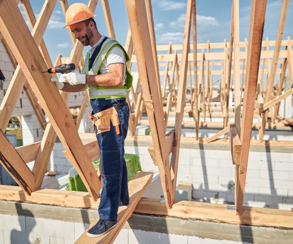 Kind bearded builder wearing helmet and uniform while using screwdriver for fixing beams