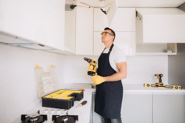 Maintenance man installing kitchen furniture in a black apron and yellow gloves using a power skrewdriver